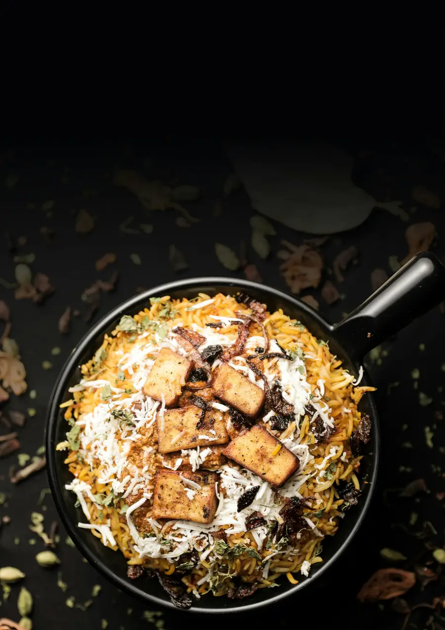 Biryani in einer schwarzen Handi-Pfanne, ringsum ganze Gewürze auf dunklem Grund.