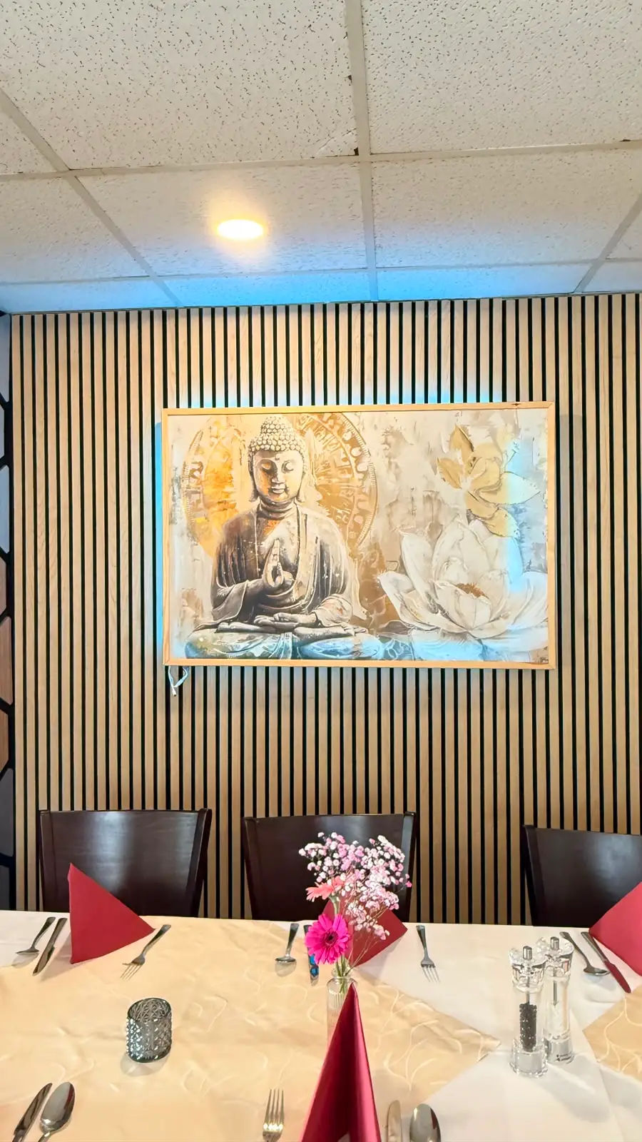 Das Buddha-Bild auf der hellen Lattenwand, darunter ein eingedeckter Tisch mit rot gefalteten Servietten und frischen Blumen.