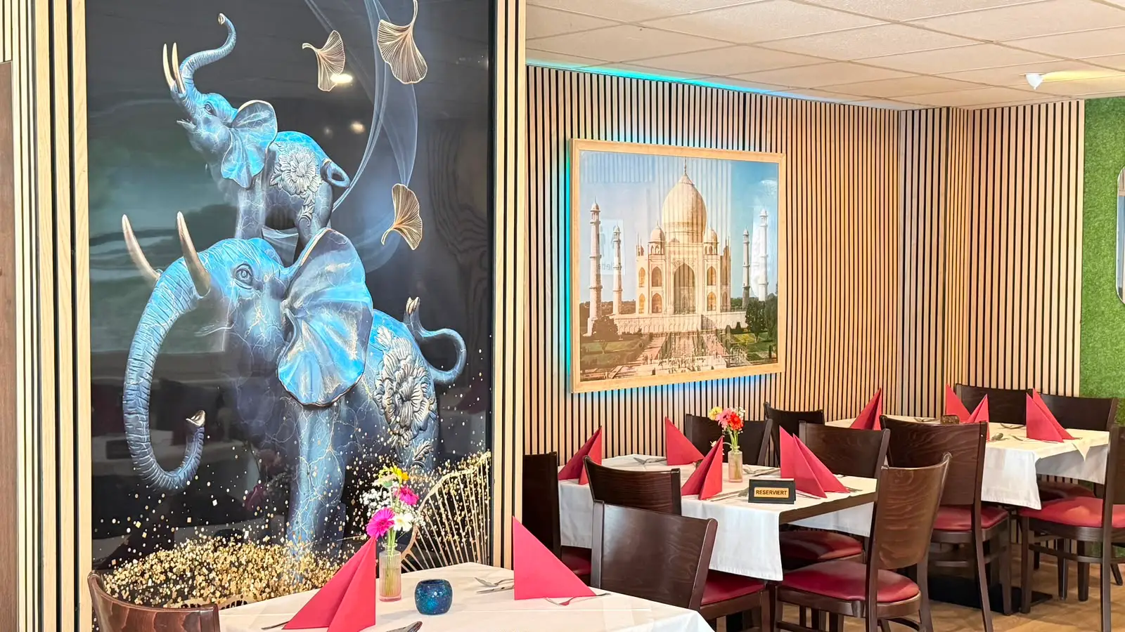 Elefanten-Wandbild und Taj-Mahal-Bild an der Lattenwand, davor die eingedeckten Tische des Gastraums.